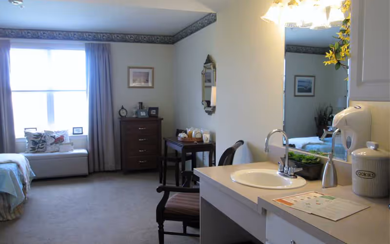 Studio-style bedroom with a window seat, dresser, small table and a sink/vanity in the foreground.