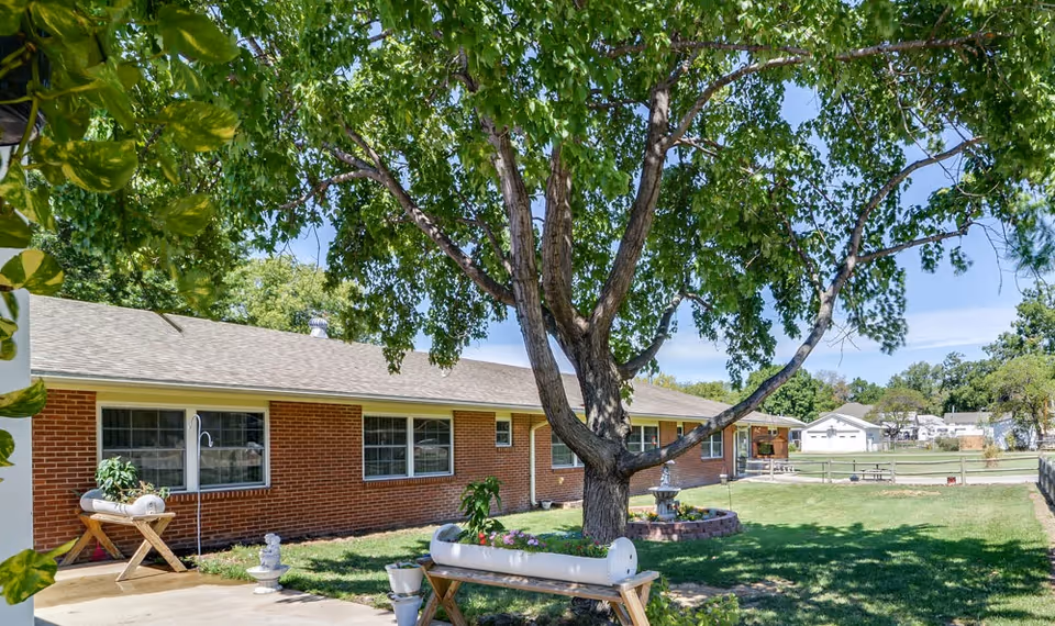 Outdoor view of a single-story brick building with multiple windows, a large tree with green leaves in the foreground, and a grassy yard with planters and a small fountain.