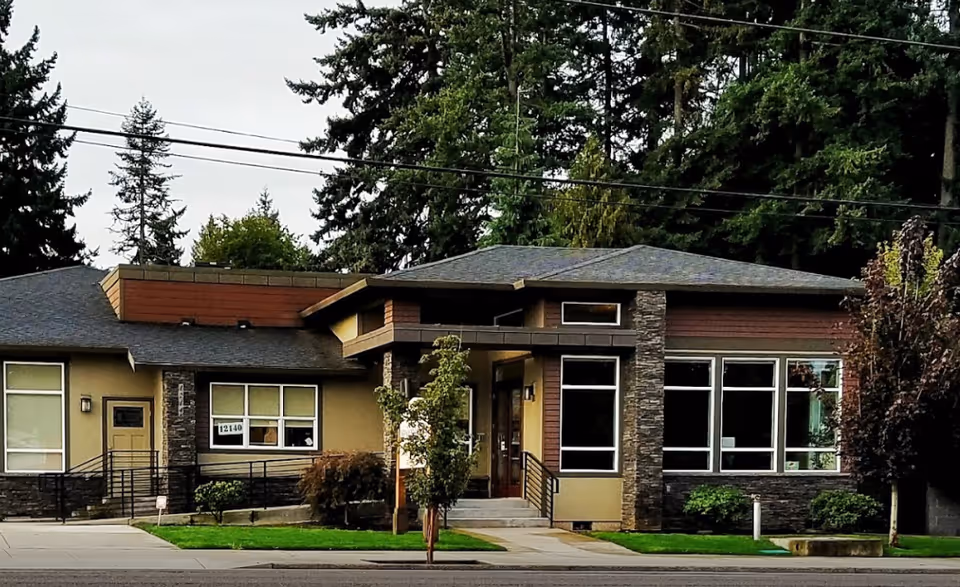 Exterior view of a single-story senior living facility building with modern architecture, featuring large windows, stone and wood accents, a small front lawn with trees and shrubs, and a sidewalk in front.