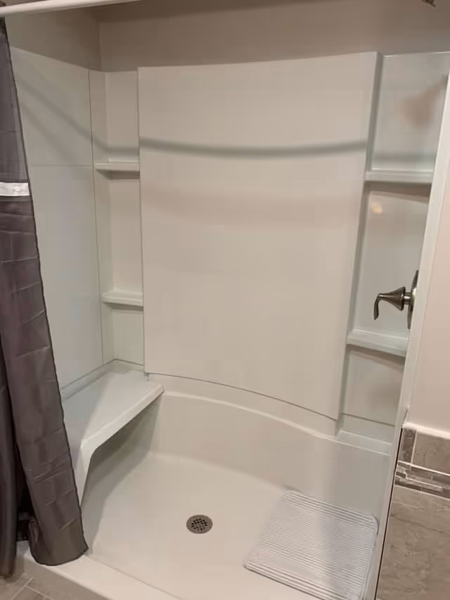 A white walk-in shower stall with a built-in corner seat and shelves, gray shower curtain, metal faucet handle, and a small bath mat.