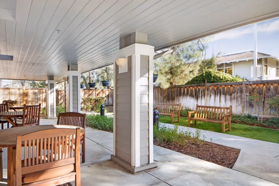 Covered outdoor patio area with wooden tables and chairs, surrounded by a garden with green grass, plants, and wooden benches. A wooden fence and trees are visible in the background under a clear sky.
