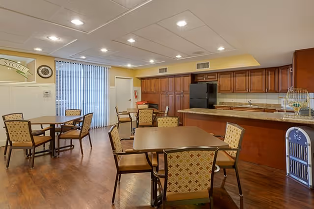 Communal dining area with several tables and upholstered chairs facing a kitchenette with wood cabinets and a refrigerator.