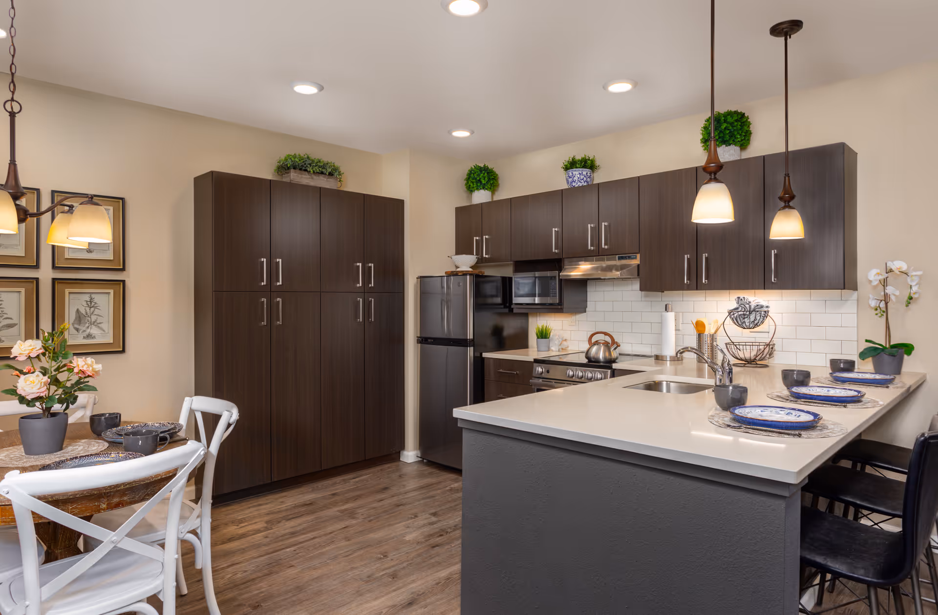 Modern kitchen and dining area in an assisted living community featuring dark wood cabinets, stainless steel refrigerator and stove, white countertops with a sink, three pendant lights hanging above the counter with place settings, a small round dining table with white chairs, decorative plants, and framed botanical prints on the wall.