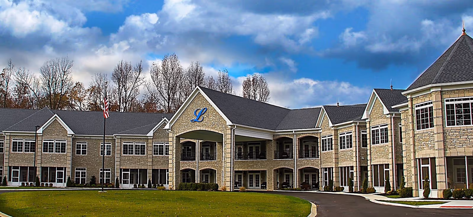 Exterior view of The Lakes of Paducah senior living facility showing a large two-story building with stone facade, multiple windows, a covered entrance with balconies above, an American flag on a flagpole, and a green lawn in front under a partly cloudy sky.