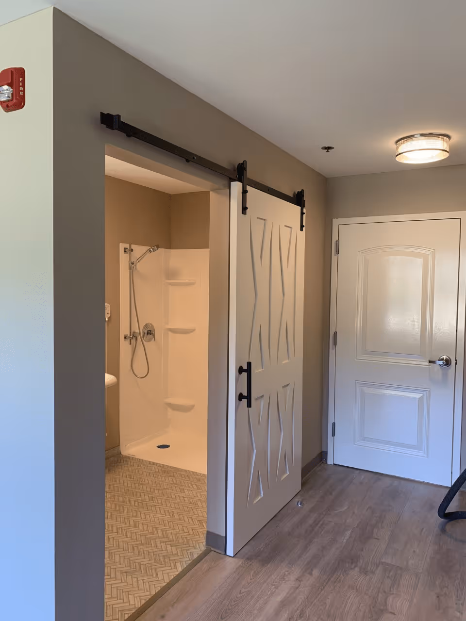Interior view of a senior living facility bathroom with a sliding barn door partially open, revealing a walk-in shower with a handheld showerhead and built-in shelves. The room has beige walls, wood flooring, and a white door with a silver handle.