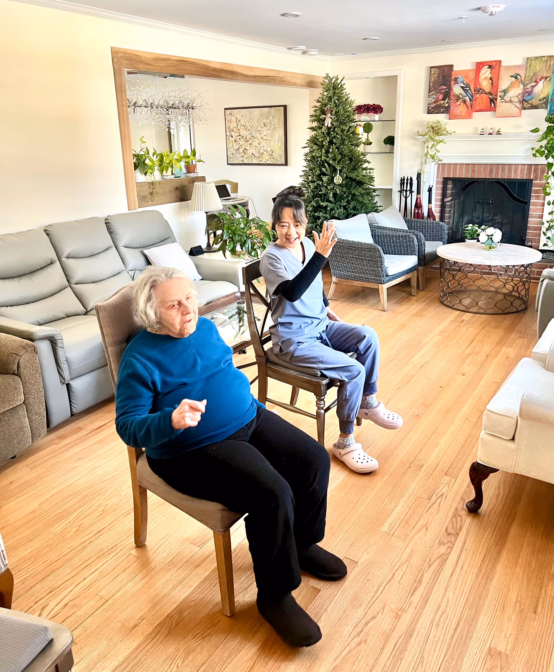 An elderly woman and a caregiver in scrubs sit on chairs in a bright assisted-living living room with a Christmas tree, fireplace, and seating area.