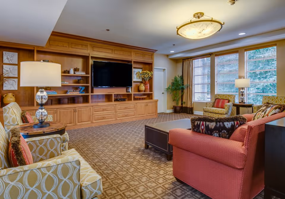 A cozy living room with patterned carpet, a large wooden entertainment center with a flat-screen TV, two patterned armchairs with red cushions, a red sofa with decorative pillows, two side tables with lamps, and large windows with blinds letting in natural light.