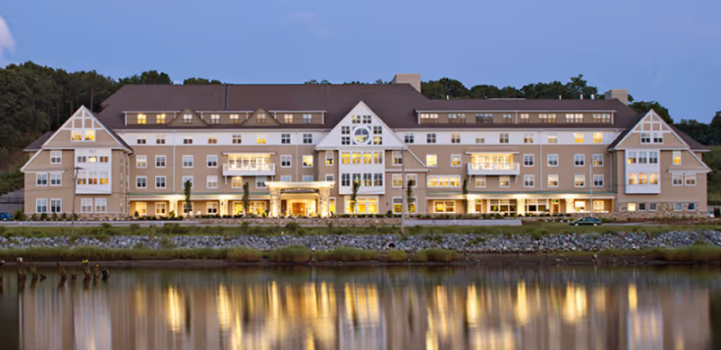 A large multi-story waterfront senior living building lit at dusk with its lights reflecting in the calm water.
