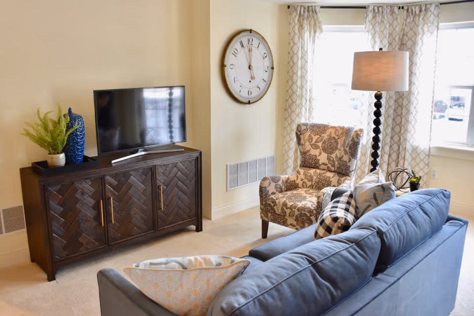 A cozy living room with a blue sofa adorned with patterned pillows, a floral armchair, a dark wooden TV stand with a flat-screen television, a tall floor lamp, a large wall clock, and windows with patterned curtains letting in natural light.