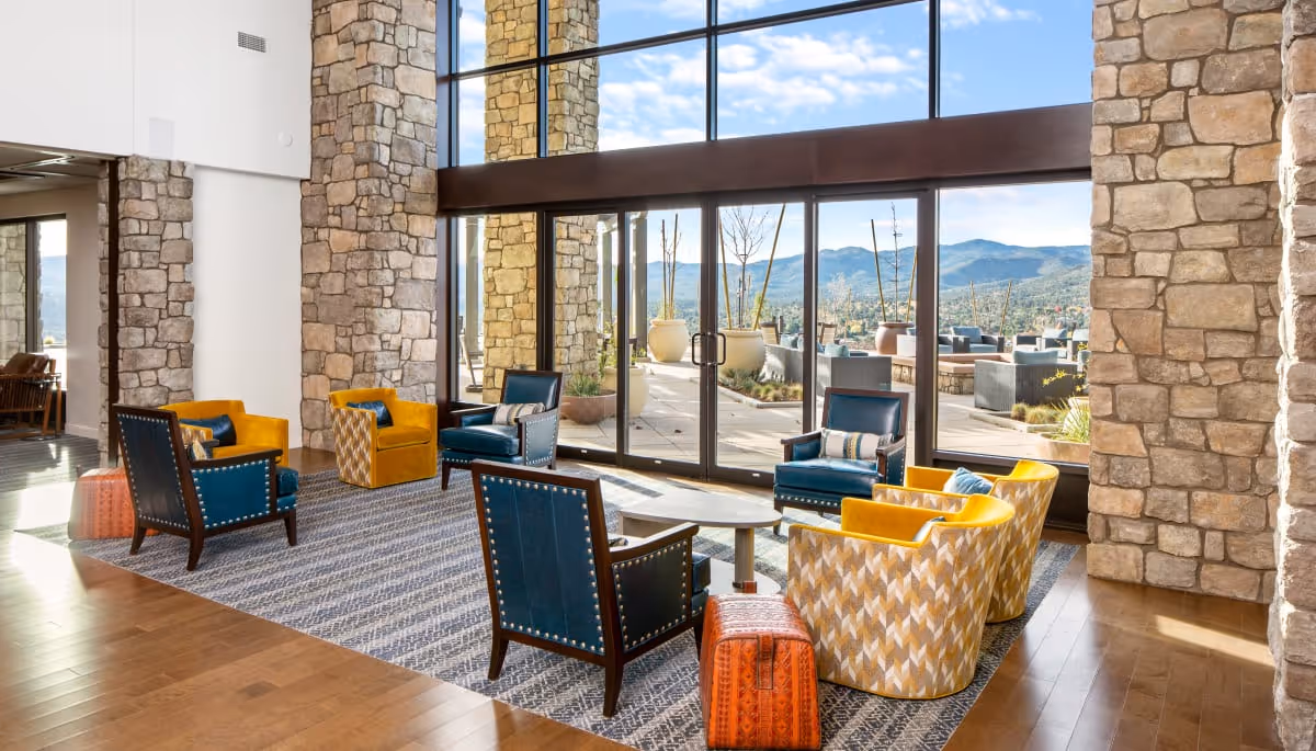 A bright and spacious living room area with large stone pillars and floor-to-ceiling windows offering a scenic mountain view. The room features a seating arrangement with blue and yellow armchairs around a small wooden coffee table, placed on a patterned rug. Outside the windows, there is a patio with additional seating and large potted plants.
