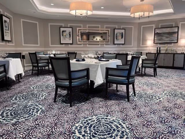 A formal dining room with round tables covered in white tablecloths, set with glassware and napkins. The room features patterned carpet, gray paneled walls with framed artwork, and two large ceiling light fixtures. There are black chairs with blue cushions around the tables.