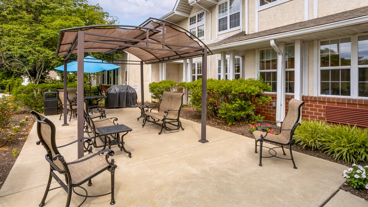 Outdoor patio area at Brookdale Northampton with metal chairs and tables under a canopy and a blue umbrella, surrounded by greenery and the exterior of the building with multiple windows.