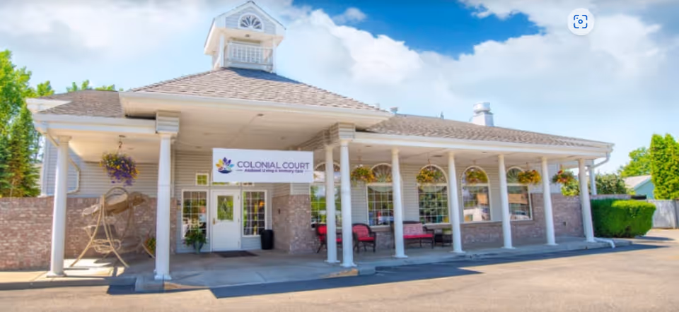 Front exterior view of Colonial Court assisted living and memory care facility with a covered entrance supported by white columns, hanging flower baskets, and outdoor seating.