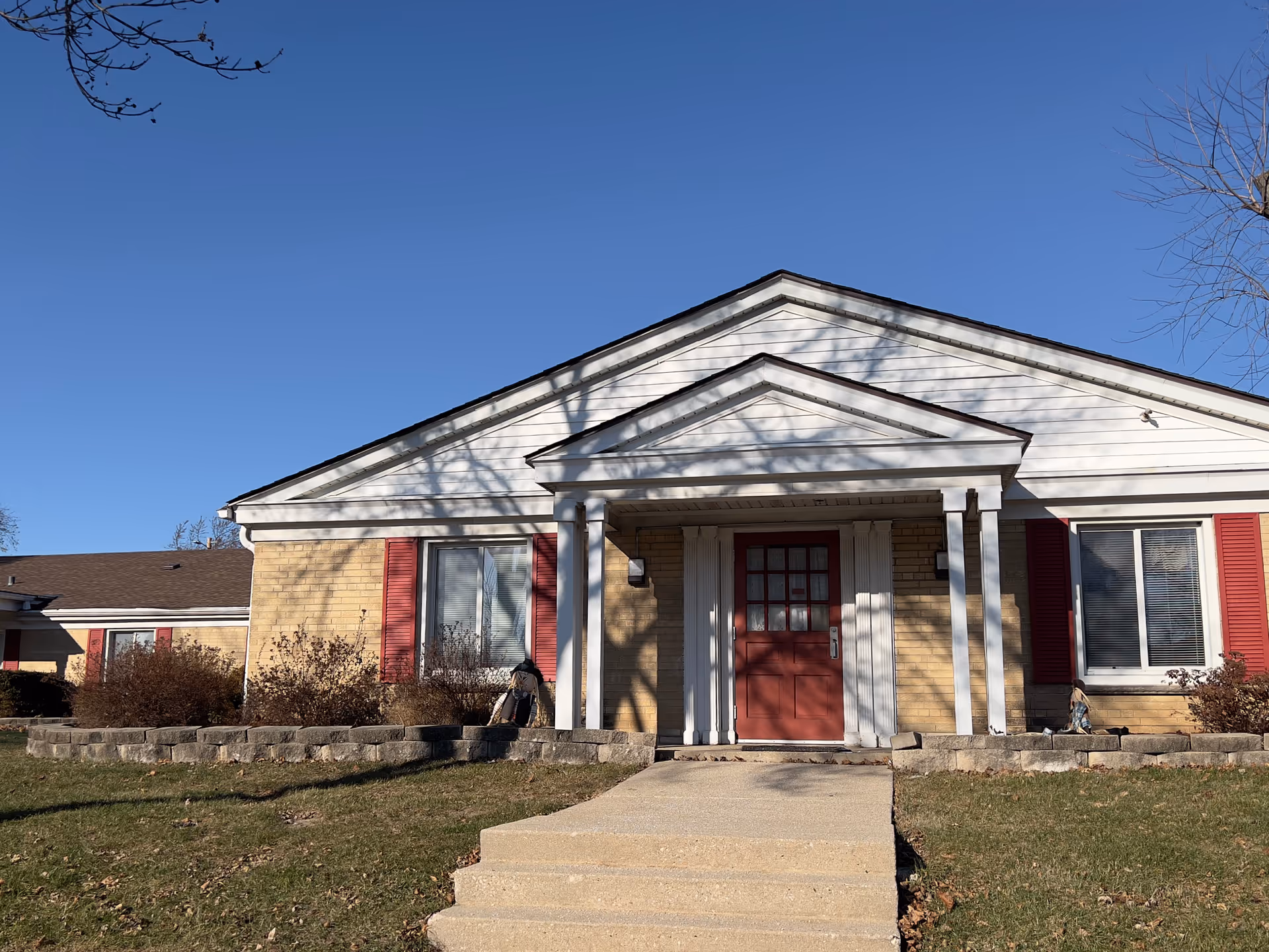 Front exterior view of a single-story building with beige brick walls, white trim, red shutters, and a red door under a small portico with white columns. There are steps leading up to the entrance and some landscaping with bushes and grass in front.