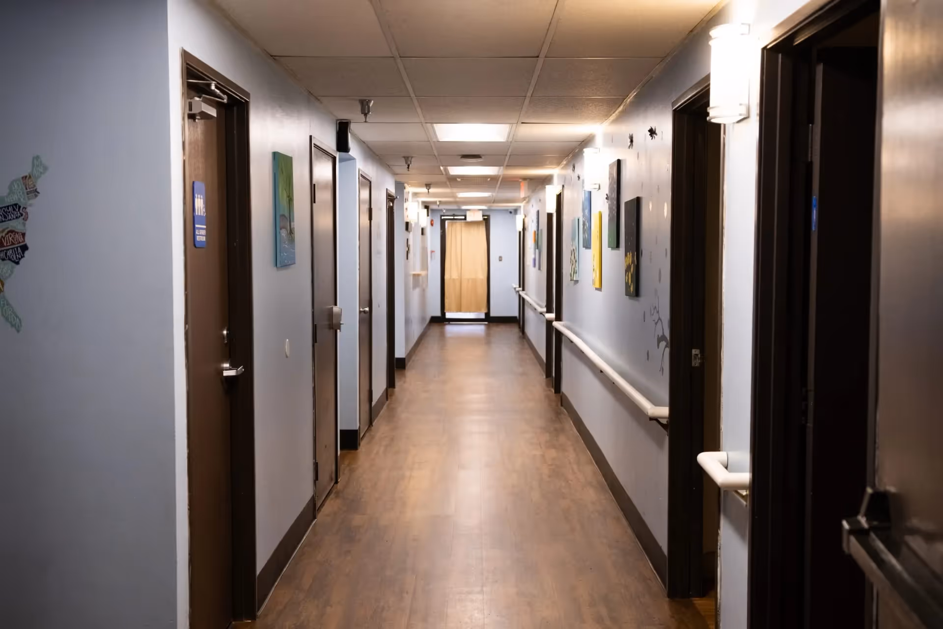 A long hallway in a senior living facility with light blue walls, wooden doors on both sides, handrails along the walls, and various colorful paintings hanging on the right wall. The floor is wooden, and the ceiling has recessed lighting panels.