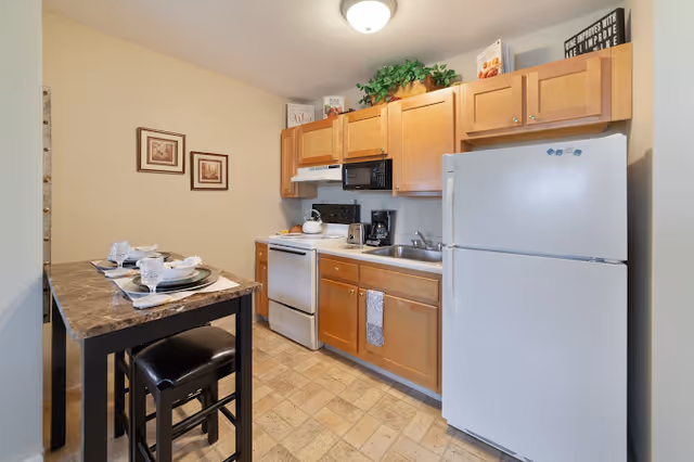 Compact kitchen with light wood cabinets, white refrigerator and stove, microwave, sink, and a small dining table set for two.