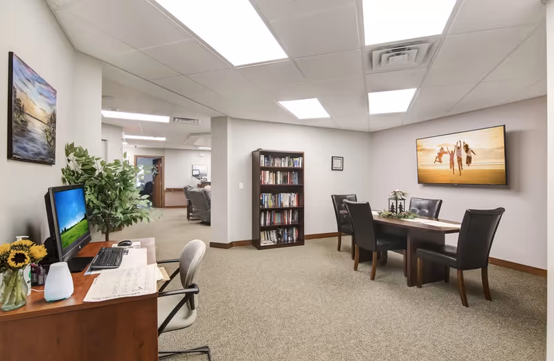 Well-lit interior common area with a desk and computer on the left, a bookshelf, and a dining-style table with four chairs beneath a wall-mounted TV.