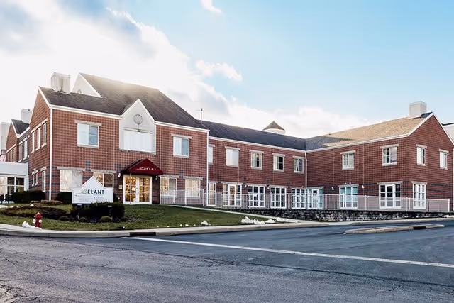 Exterior view of a two-story brick senior living facility building with multiple windows and a red awning over the entrance. There is a sign on the lawn near the entrance that reads 'Elant'. The sky is partly cloudy with sunlight illuminating the building.