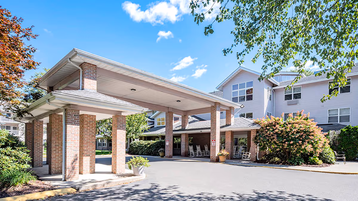 Exterior view of Holiday Quail Run Estates showing a covered entrance supported by brick columns, surrounded by greenery and trees under a blue sky with some clouds.