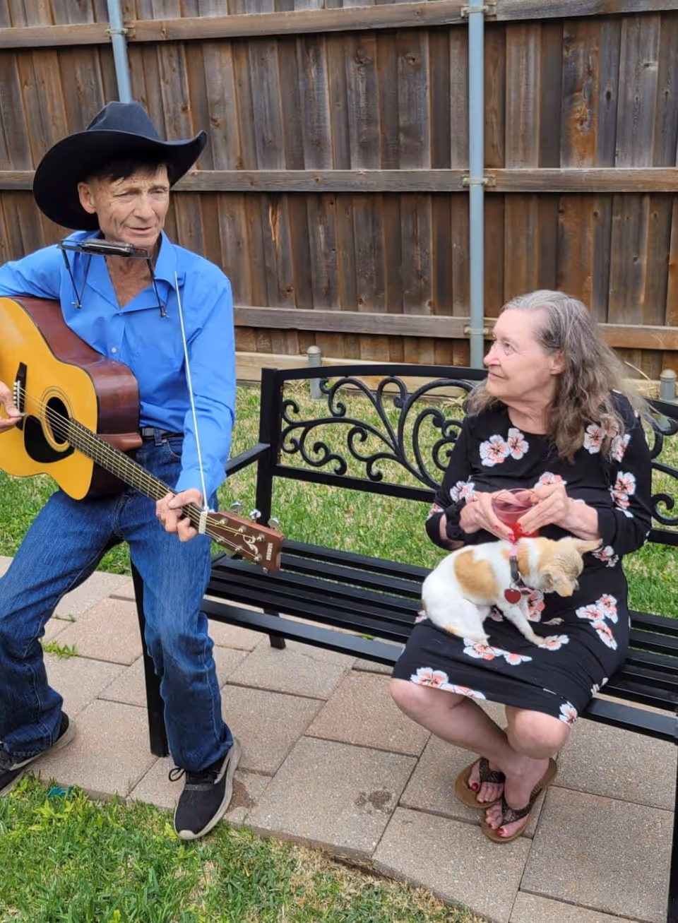 An elderly man wearing a black cowboy hat and blue shirt is playing an acoustic guitar and harmonica while sitting on a bench outdoors. An elderly woman in a black floral dress is sitting on the same bench holding a small dog on her lap, looking at the man. They are in a fenced backyard with grass and a paved area.