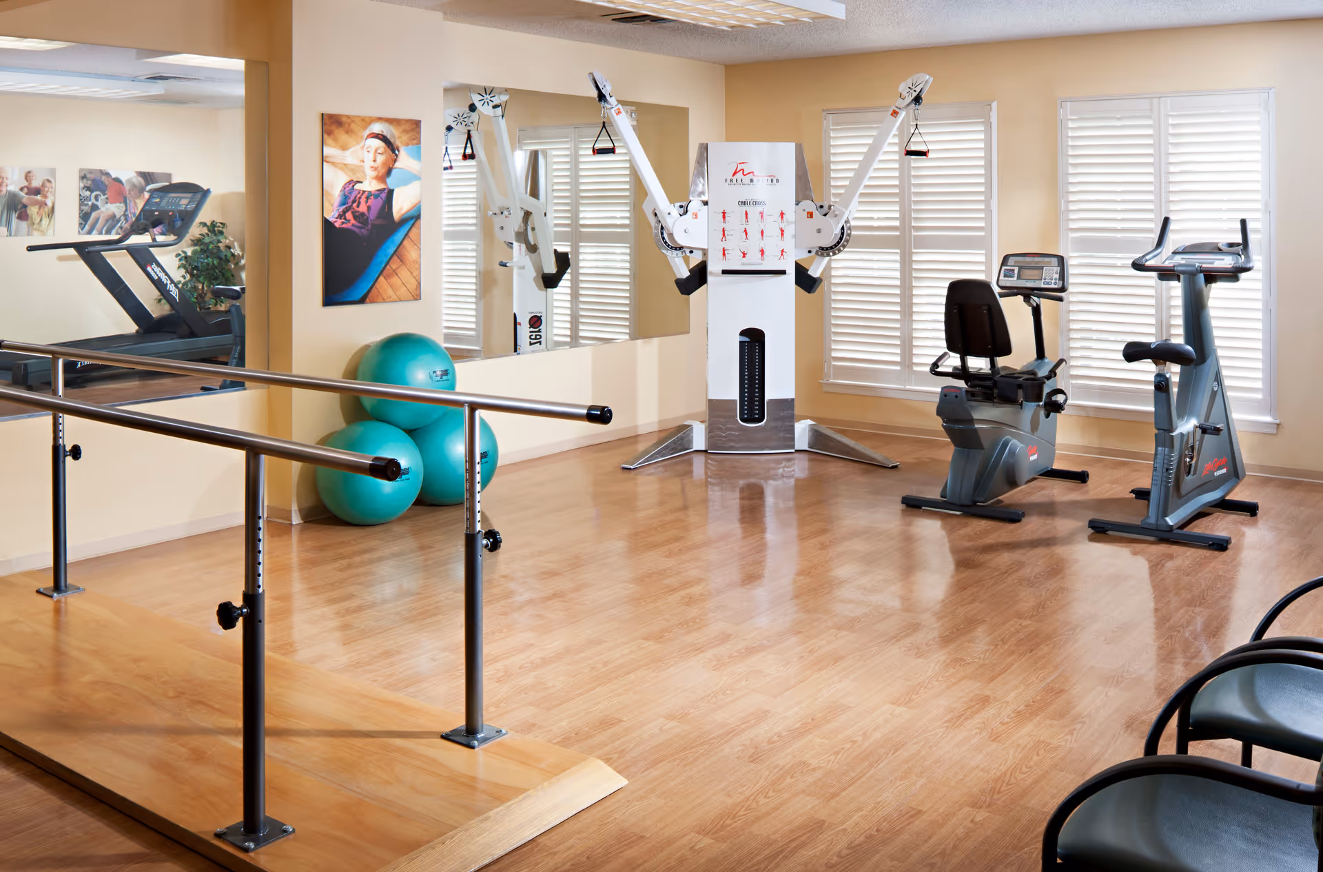 A bright exercise room with wooden flooring featuring fitness equipment including a treadmill, stationary bikes, a multi-functional weight machine, and three green exercise balls. There is a ballet barre in the foreground and large mirrors on the wall reflecting the equipment. The room has beige walls, white window shutters, and framed photos on the walls.