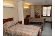 A shared bedroom in a senior living facility with two single beds, each with patterned bedspreads. There is a curtain divider between the beds, a wooden nightstand with a lamp, a crucifix on the wall, a window with blinds, and a small cabinet with items on top.