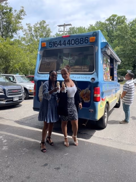 Two women standing in front of a blue ice cream truck holding ice cream cones in a parking lot with trees and other parked cars in the background. A man is standing to the right side near the truck.