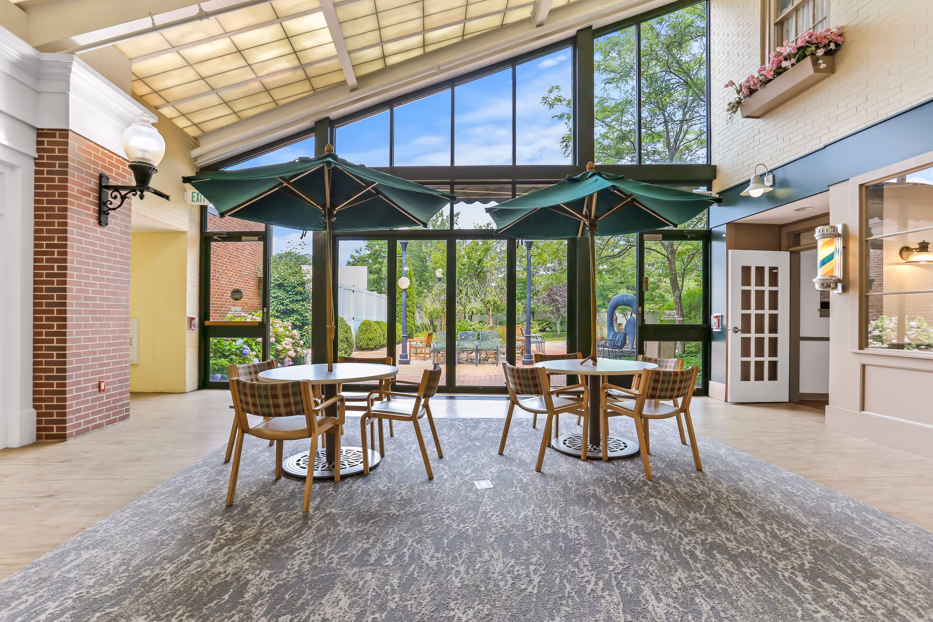 Indoor seating area with two round tables, each shaded by a large green umbrella. Each table is surrounded by four wooden chairs with plaid cushions. The area has a large glass wall and door leading to an outdoor patio with greenery and trees visible outside. The ceiling is high with a translucent panel allowing natural light. There is a brick wall on the left and a white wall with a flower box on the right.
