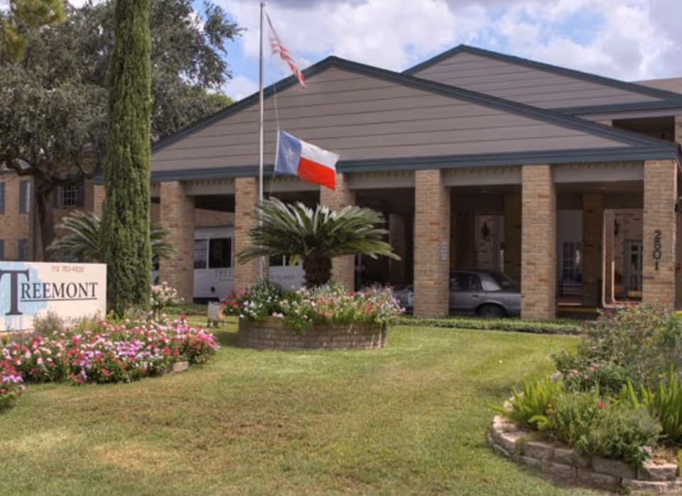 Front exterior view of Treemont Senior Living facility with a covered entrance supported by brick columns, a flagpole displaying the American and Texas flags, and landscaped flower beds and greenery in the foreground.