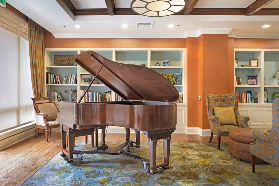 A cozy living room area featuring a grand piano with its lid open, surrounded by upholstered chairs and a patterned rug. Behind the piano, there are built-in white bookshelves filled with books and decorative items. The room has warm orange walls, wooden flooring, and a large window with curtains allowing natural light to enter.