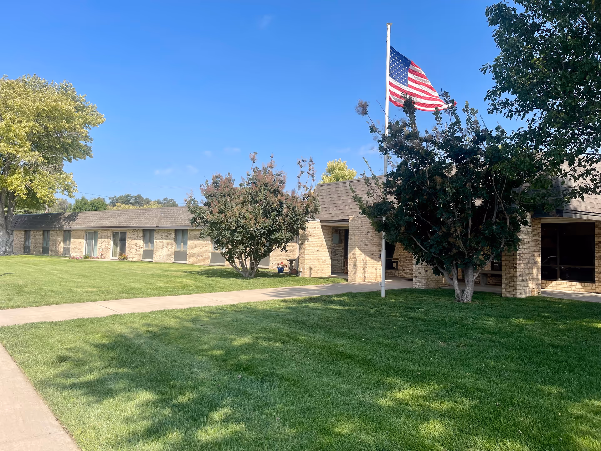 Exterior view of a single-story brick building with a shingled roof, surrounded by green grass and trees. An American flag is flying on a flagpole near the entrance under a clear blue sky.