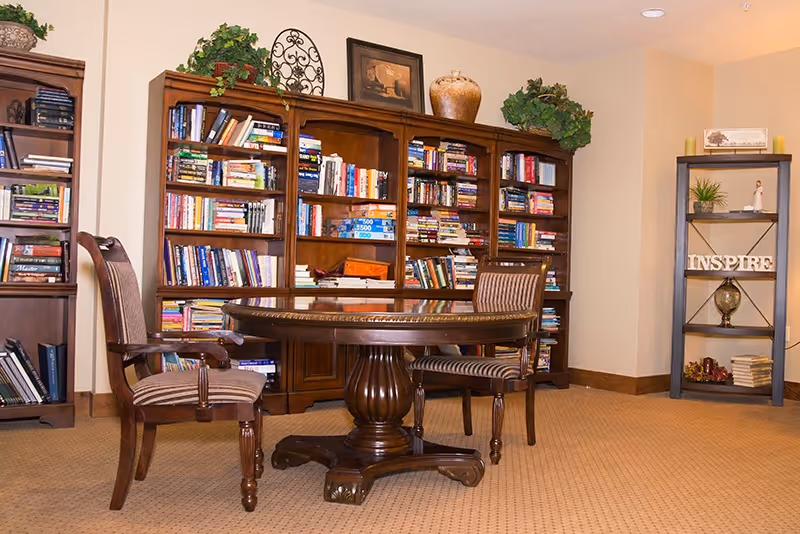 A cozy interior room featuring a round wooden table with two striped upholstered chairs. Behind the table are large wooden bookshelves filled with books and decorative items. To the right, there is a smaller shelving unit with plants, books, and a decorative sign that reads 'INSPIRE'. The room has beige walls and carpeted flooring.
