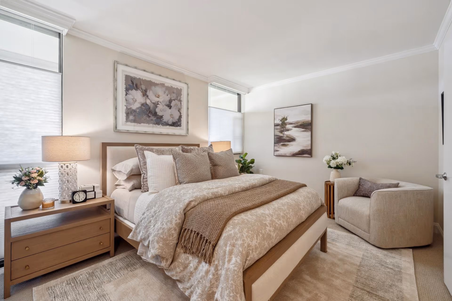 A cozy bedroom with a neatly made bed featuring multiple pillows and a beige patterned comforter. There is a wooden nightstand with a lamp, a clock, a small stack of books, and a vase with flowers. The room has two large windows with white blinds, a beige armchair with a decorative pillow, a small wooden side table with a vase of flowers, and two framed paintings on the walls. The color scheme is neutral with soft beige and brown tones.
