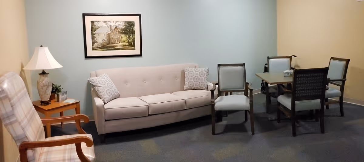 A cozy senior living community room featuring a beige sofa with two patterned pillows, a wooden armchair with plaid upholstery, a wooden side table with a lamp and small plant, and a square table surrounded by four chairs. The walls are painted in soft beige and light blue, and a framed picture of a house hangs above the sofa.