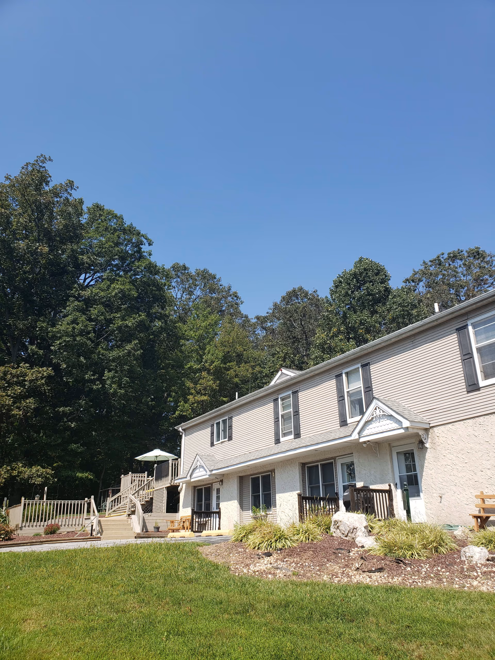 Exterior view of a two-story residential building with beige siding and white trim, surrounded by green grass and trees under a clear blue sky. There is a wooden deck with an umbrella and outdoor seating on the left side.
