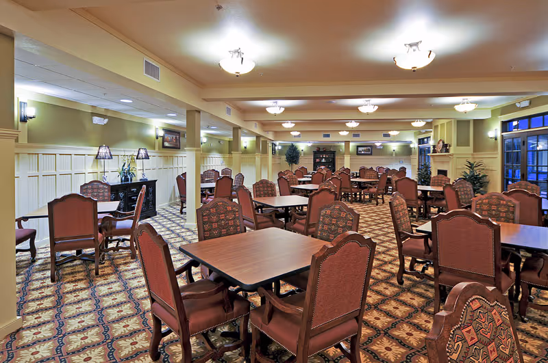 Spacious, warmly lit dining room with multiple square tables and upholstered chairs arranged on a patterned carpet.