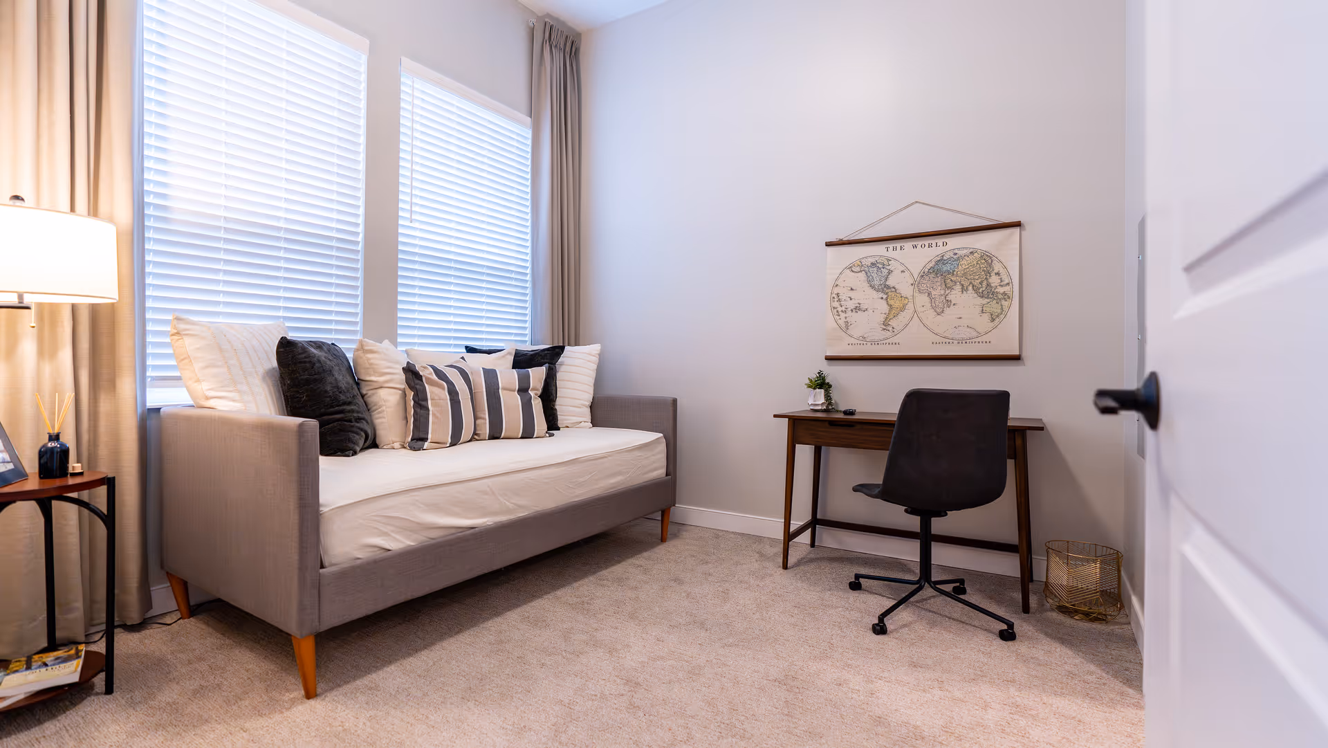 A cozy room with a gray daybed adorned with multiple pillows in white, black, and striped patterns. Next to the daybed is a small side table with a lamp, a reed diffuser, and a framed photo. Across from the daybed is a wooden desk with a black swivel chair, a small plant, and a remote control. Above the desk hangs a vintage-style world map. The room has beige carpet, light-colored walls, and two large windows with white blinds and beige curtains.
