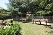 Outdoor garden area with raised wooden garden beds, green grass, plants, and trees. A wooden fence and a building are visible in the background. A patriotic banner is hanging on the fence.