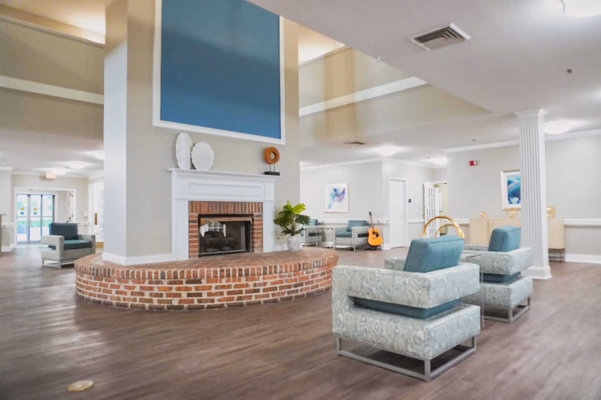 A spacious and well-lit assisted living facility common area with a large brick fireplace in the center. The room features modern armchairs with patterned upholstery and teal cushions, a guitar resting against the wall, decorative art pieces on the walls, and a potted plant on the fireplace hearth. The flooring is wood-style, and the ceiling has recessed lighting and ventilation.