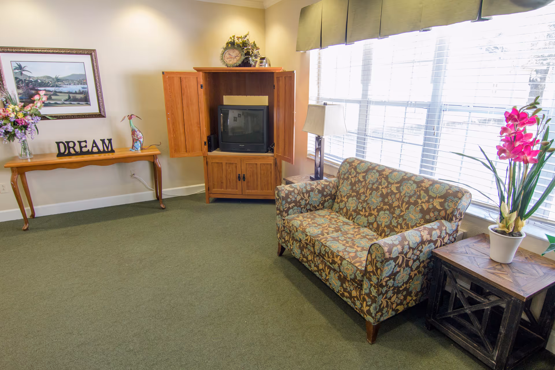 A cozy living room area with a floral patterned loveseat next to a wooden side table holding a potted plant with pink flowers. There is a wooden cabinet with open doors revealing a television inside. A table against the wall holds a vase of flowers, a decorative bird, and a sign that says 'DREAM'. A framed landscape painting hangs above the table. Large windows with blinds allow natural light to fill the room.