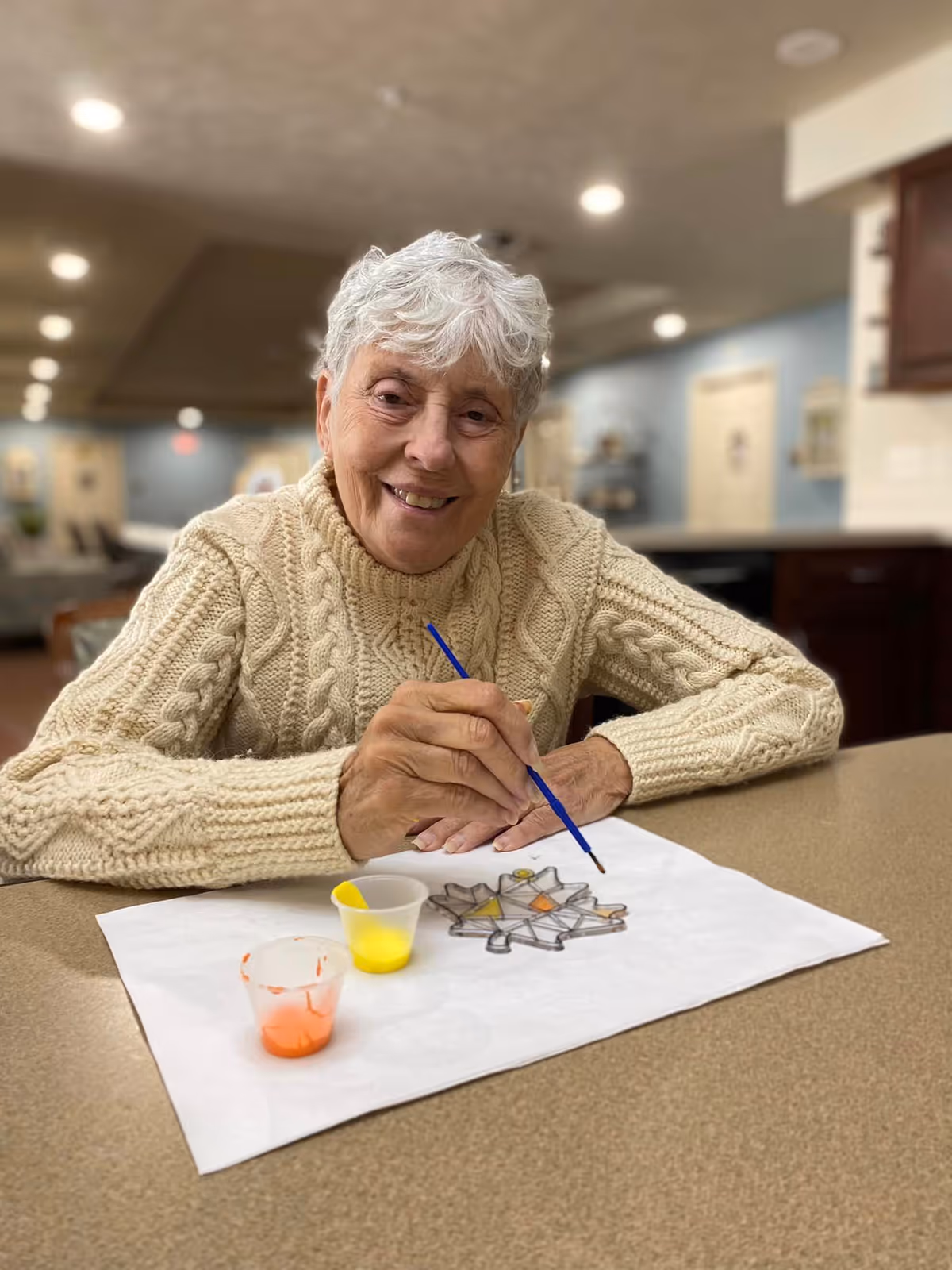 An elderly woman with short white hair wearing a cream-colored cable knit sweater is sitting at a table in a well-lit room, smiling at the camera while painting a stained glass design on a piece of paper. There are small cups of yellow and orange paint on the table in front of her.