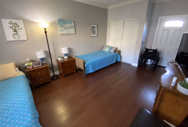 A bedroom with two single beds covered in blue bedspreads, each with pillows. Between the beds are two wooden nightstands with table lamps and decorative items. A tall floor lamp is also present. The room has wooden flooring, light gray walls with three framed pictures, a white closet door, a wheelchair near a white entrance door, and a wooden dresser with a TV on top.