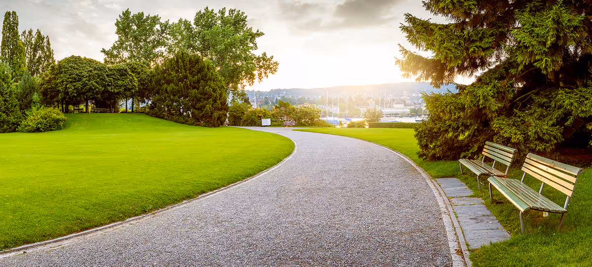 A winding gravel path through a well-maintained green park with lush trees and bushes on either side. Two wooden benches are placed on the right side near the path. In the background, there is a view of a marina with boats and a setting sun casting a warm glow over the scene.