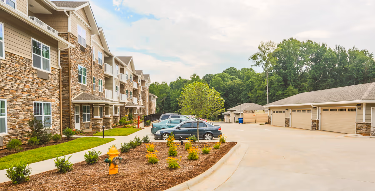Exterior view of a multi-story senior living facility with stone and beige siding, a landscaped area with small bushes and a fire hydrant, a parking lot with several cars, and a separate building with multiple garage doors. Trees and a partly cloudy sky are visible in the background.