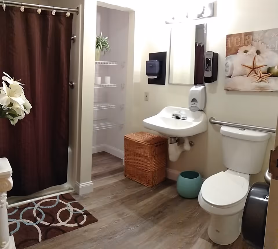 A clean bathroom with a white toilet, a wall-mounted sink, a wicker laundry basket, and a shower with a dark brown curtain. There is a small green trash bin on the floor, a wall-mounted soap dispenser, a paper towel dispenser, a mirror, and a decorative picture with starfish and towels on the wall. The floor is wood-style laminate, and there is a patterned rug in front of the shower.