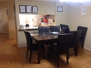 Interior view of a dining area in an assisted living facility featuring a rectangular dining table with six black chairs. The table has a dark marble-like surface with some papers and a pen on it. Behind the table is a pass-through window to the kitchen, with some kitchen items visible including a microwave and a stove. The walls are light-colored and there are framed certificates or documents hanging above the pass-through window.
