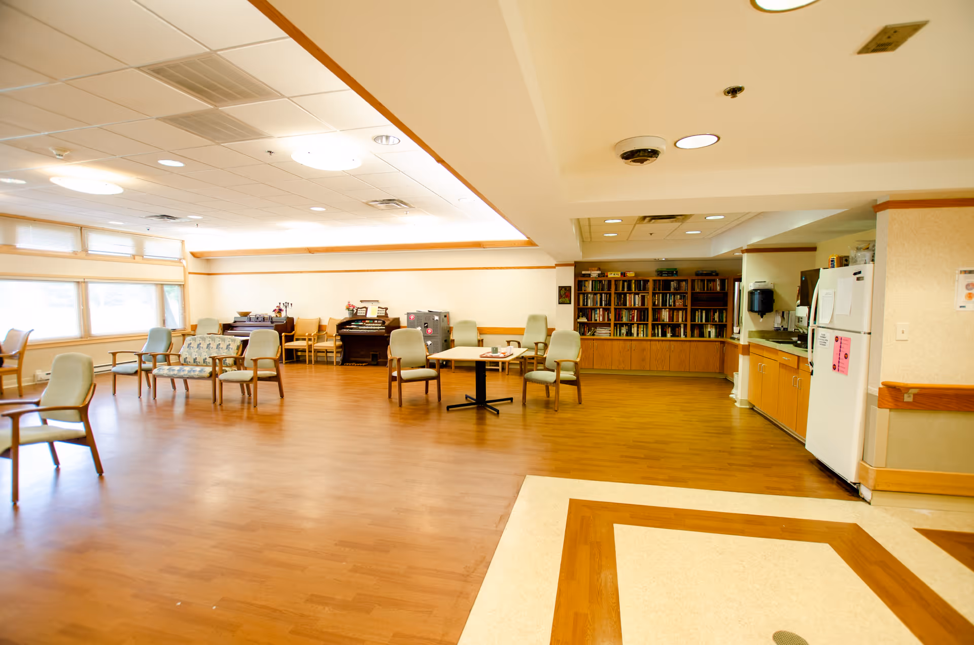 Spacious communal room in a senior facility with chairs, tables, bookshelves, and a kitchenette.
