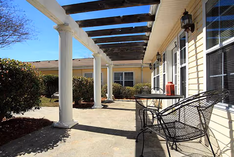 Outdoor patio area at Pinewood Square featuring a concrete floor, white columns supporting a pergola, black metal chairs and a table with a red lantern, surrounded by bushes and windows of the building.