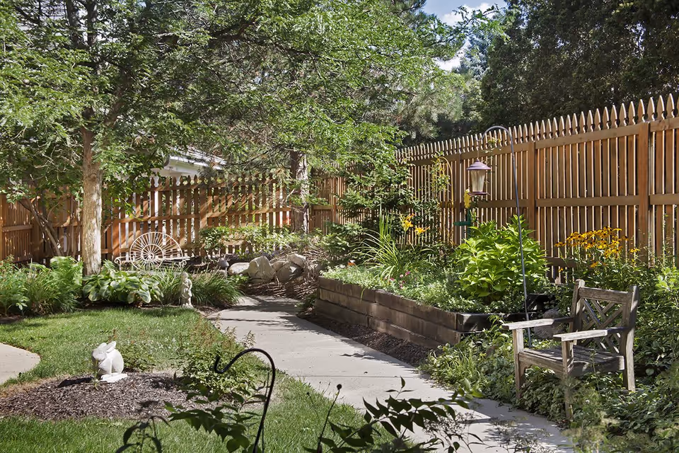 Sunlit fenced garden with a paved path, raised flower beds, lush plants, a wooden bench and decorative ornaments.