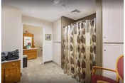 Interior view of a senior living facility bathroom area with a patterned shower curtain, cabinet sink, and adjacent dressing area.